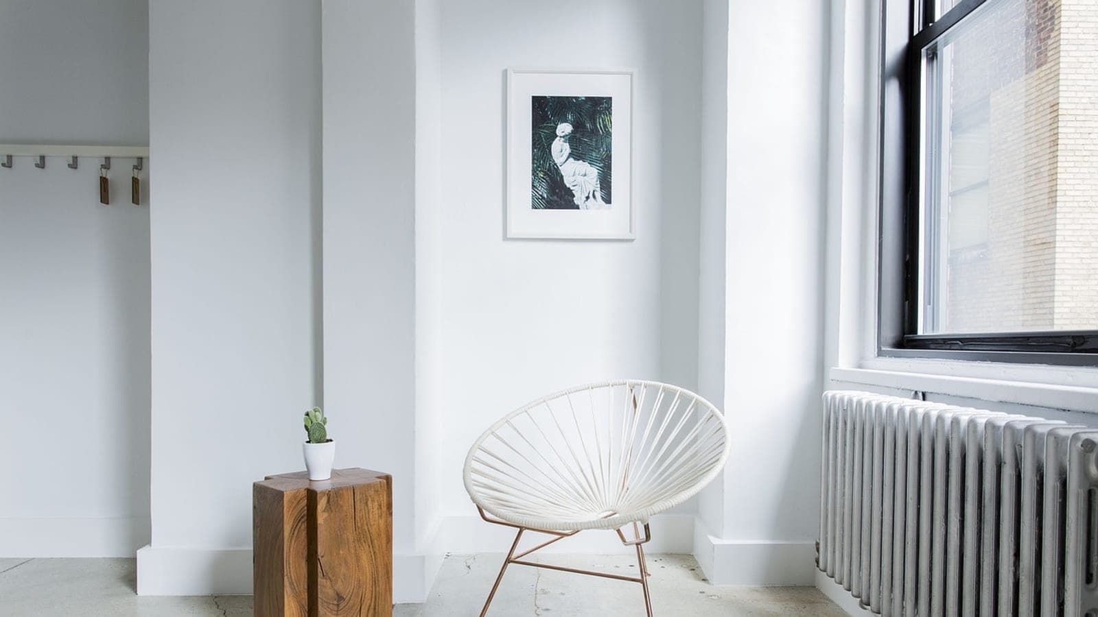 A white room with a tall radiator under a large window and a small potted plant on a wooden stool across the floor